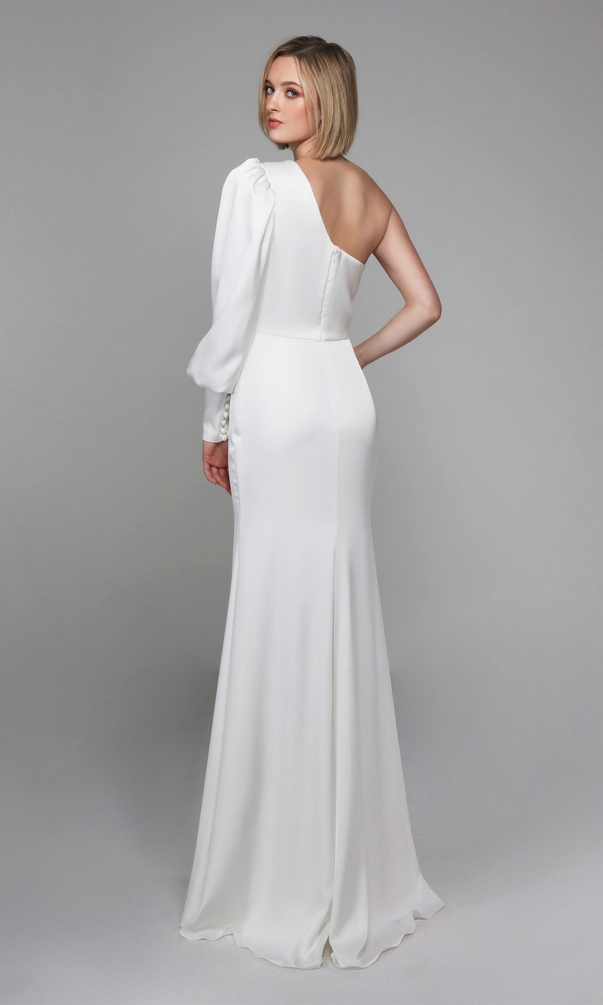 A woman faces away, wearing the Alyce 7065 long white dress—an elegant fitted one-shoulder evening gown with a single long sleeve and sleek silhouette—against a plain gray background.